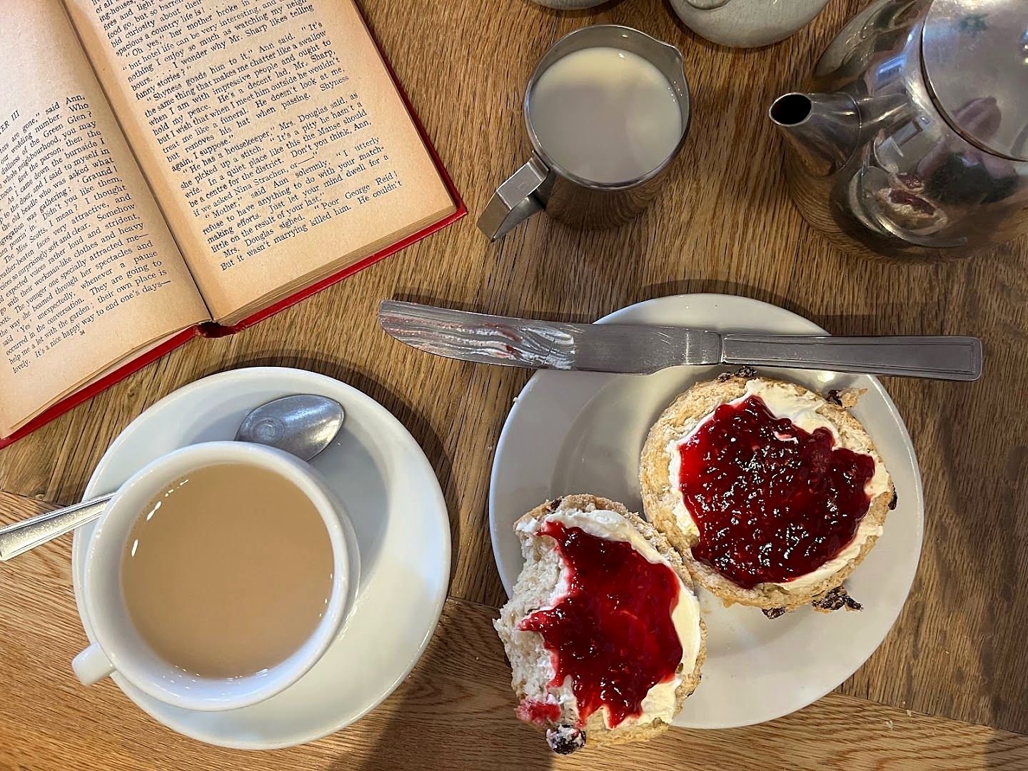 Two halved scones spread with red jam, a cup of tea, small metal teapots and an open book on a wooden table
