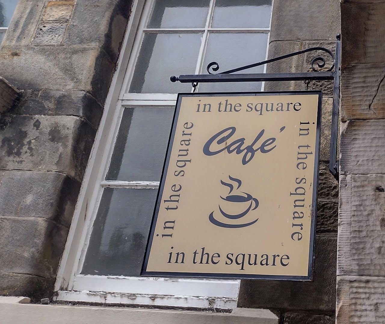 The café’s hanging sign on a stone wall: a cream square with a coffee cup, the word Café, and ‘in the square’ running round its edges
