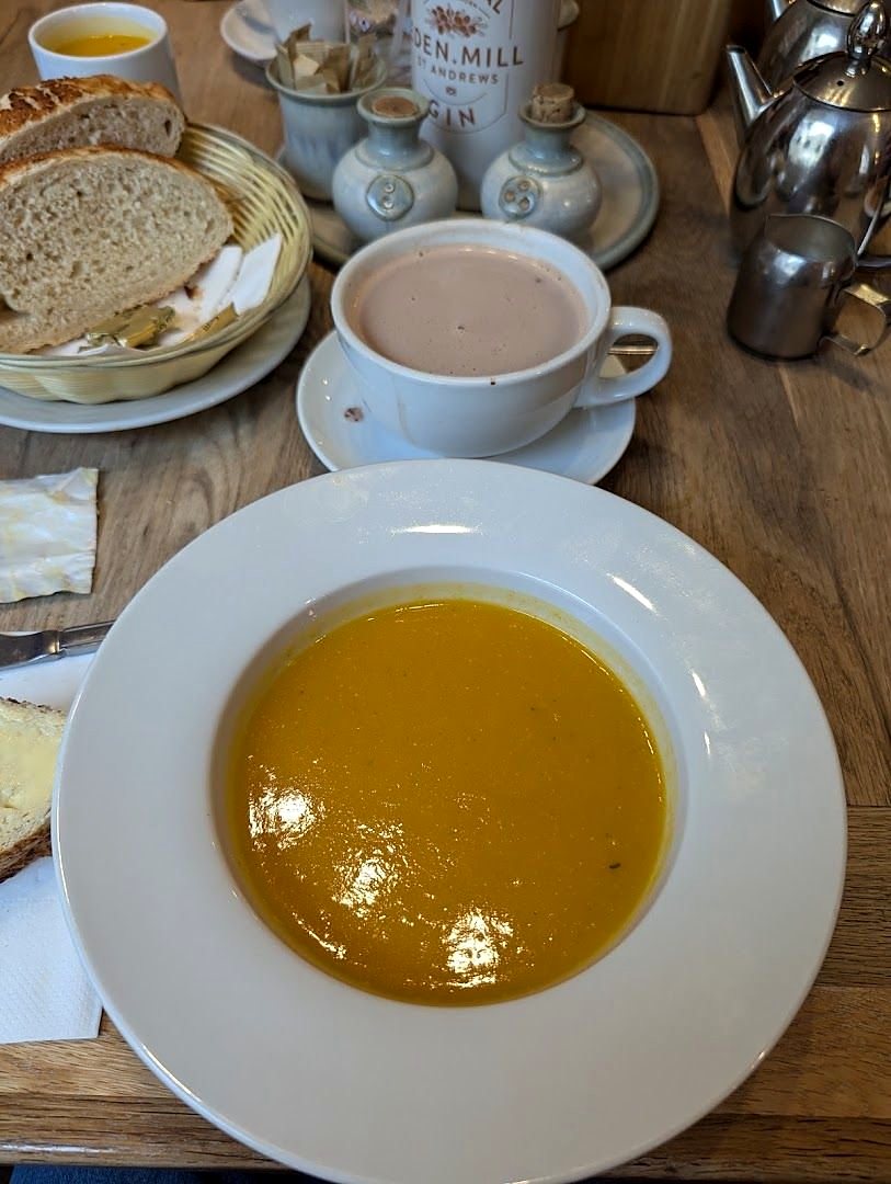 A bowl of soup on a wooden table, with a basket of sliced bread and a hot drink